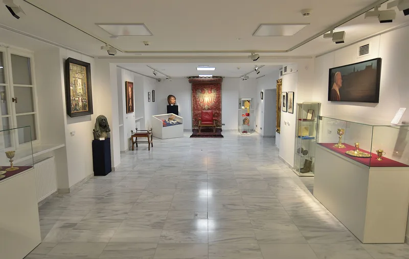 Sala na parterze Muzeum Archidiecezji Warszawskiej z gablotami liturgicznymi, portretami biskupów i czerwonym fotelem