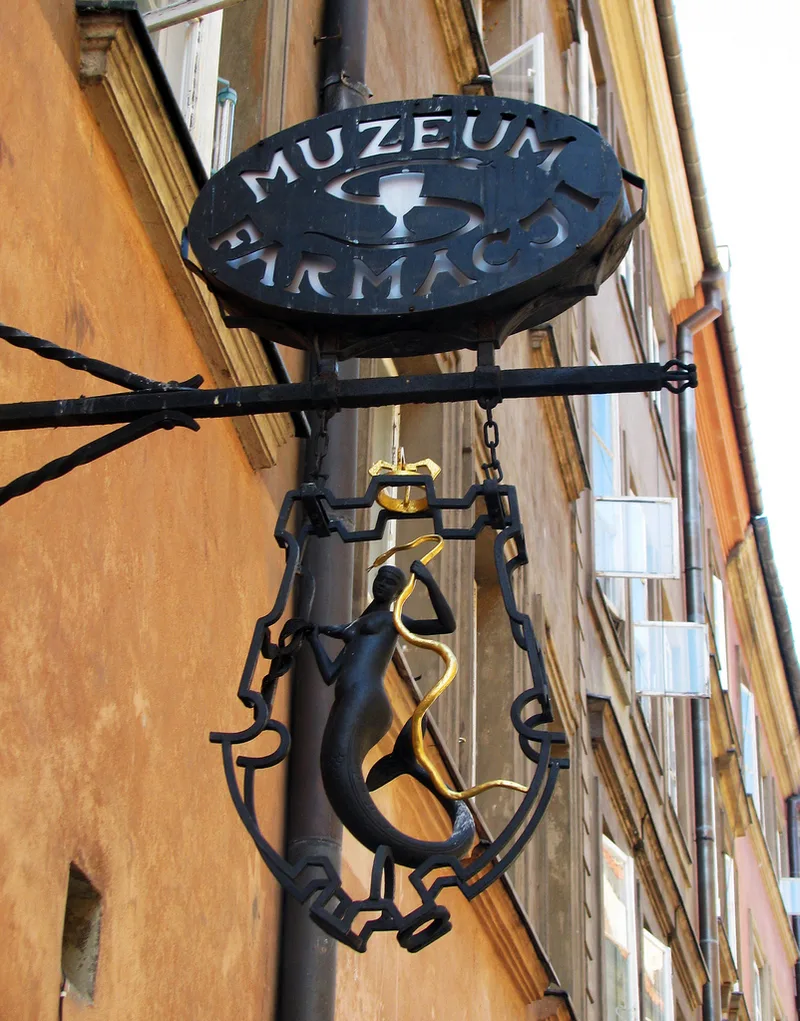 Zabytkowy żelazny szyld Muzeum Farmacji w kształcie warszawskiej syrenki, zawieszony nad wejściem na ulicy Piwnej