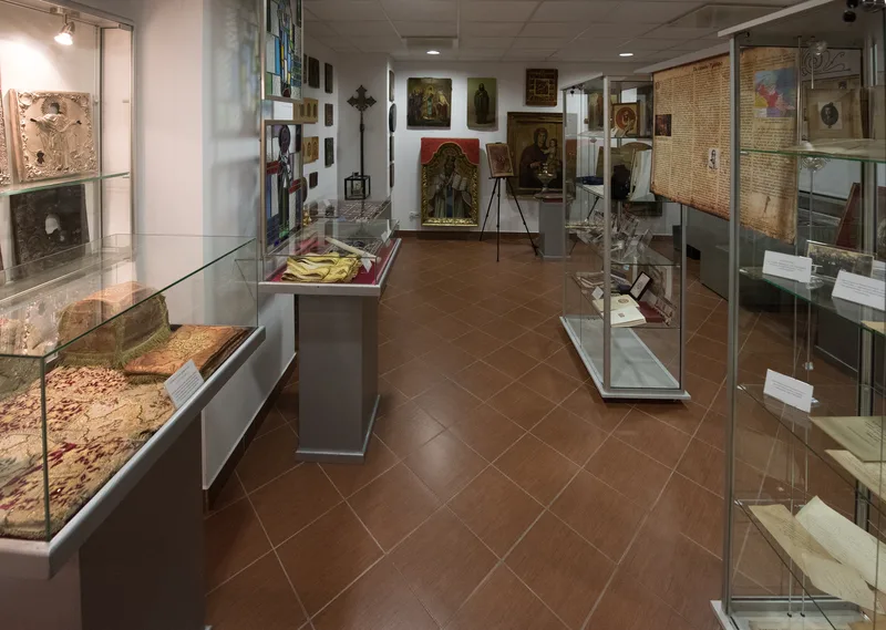 Ściana z ikonami i obrazami sakralnymi obok gabloty z tkaninami liturgicznymi